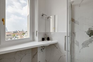 A modern bathroom with a white marble sink, a glass shower enclosure, and a large window offering a city view. - Cozy Penthouse With Stunning View Balcony (Milan)