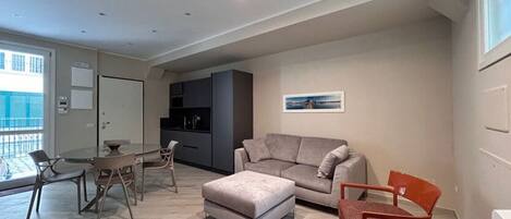 A modern living room with a grey sofa, a dining table with chairs, and a kitchen area with dark cabinets.