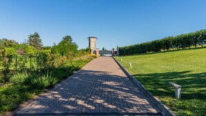 Un chemin pavé mène à un bâtiment doté d’une structure ressemblant à une tour, qui est entouré de verdure et surplombé par un ciel dégagé.