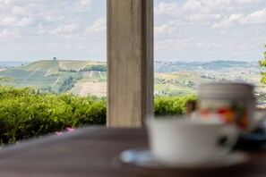 Una mesa con una taza y platillo en primer plano, un poste de madera, y una vista de colinas y campos ondulados al fondo.