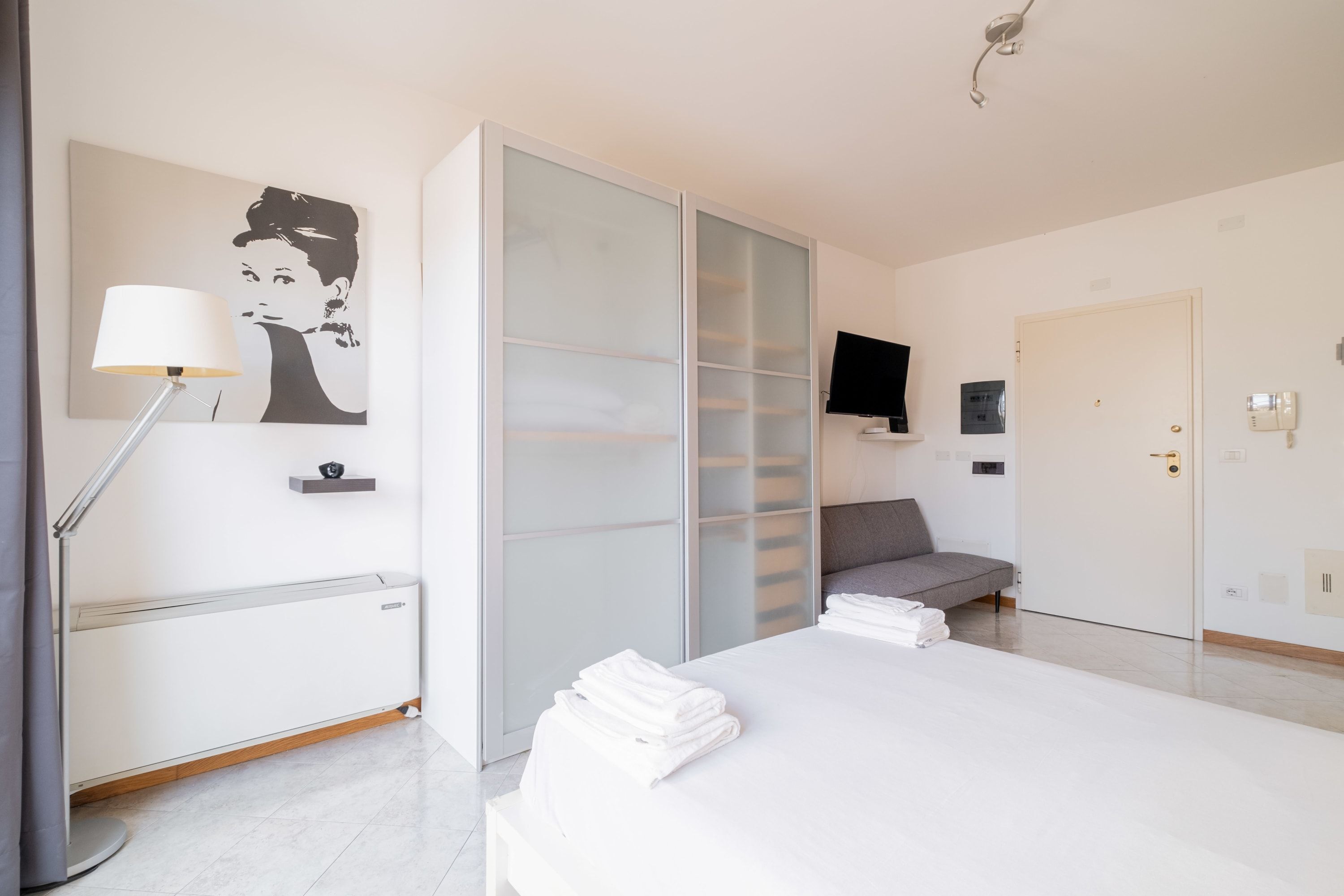Ein modernes Schlafzimmer mit Bett, Nachttischlampe, Fernseher, Schiebetürenschrank und einer Tür.