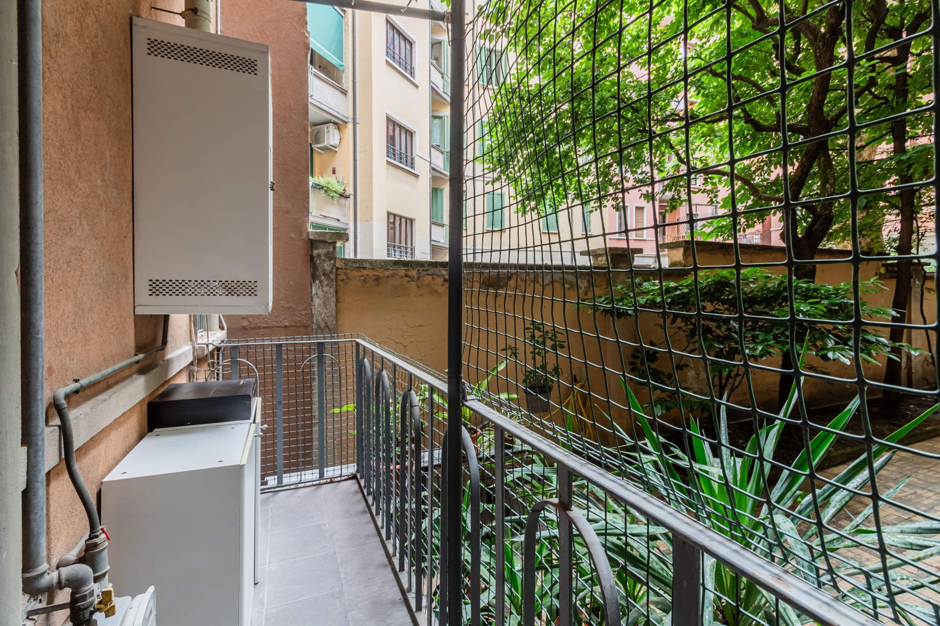 A balcony with a metal railing, a utility box, and a view of a building with balconies and greenery.