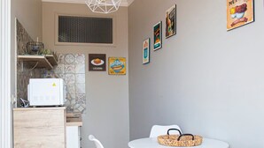Una cocina compacta con microondas, un refrigerador pequeño y un gabinete de madera. Hay una mesa redonda con cuatro sillas blancas y una cesta tejida encima. Las paredes están decoradas con cuadros enmarcados y un cartel que dice 'Lasagna'.