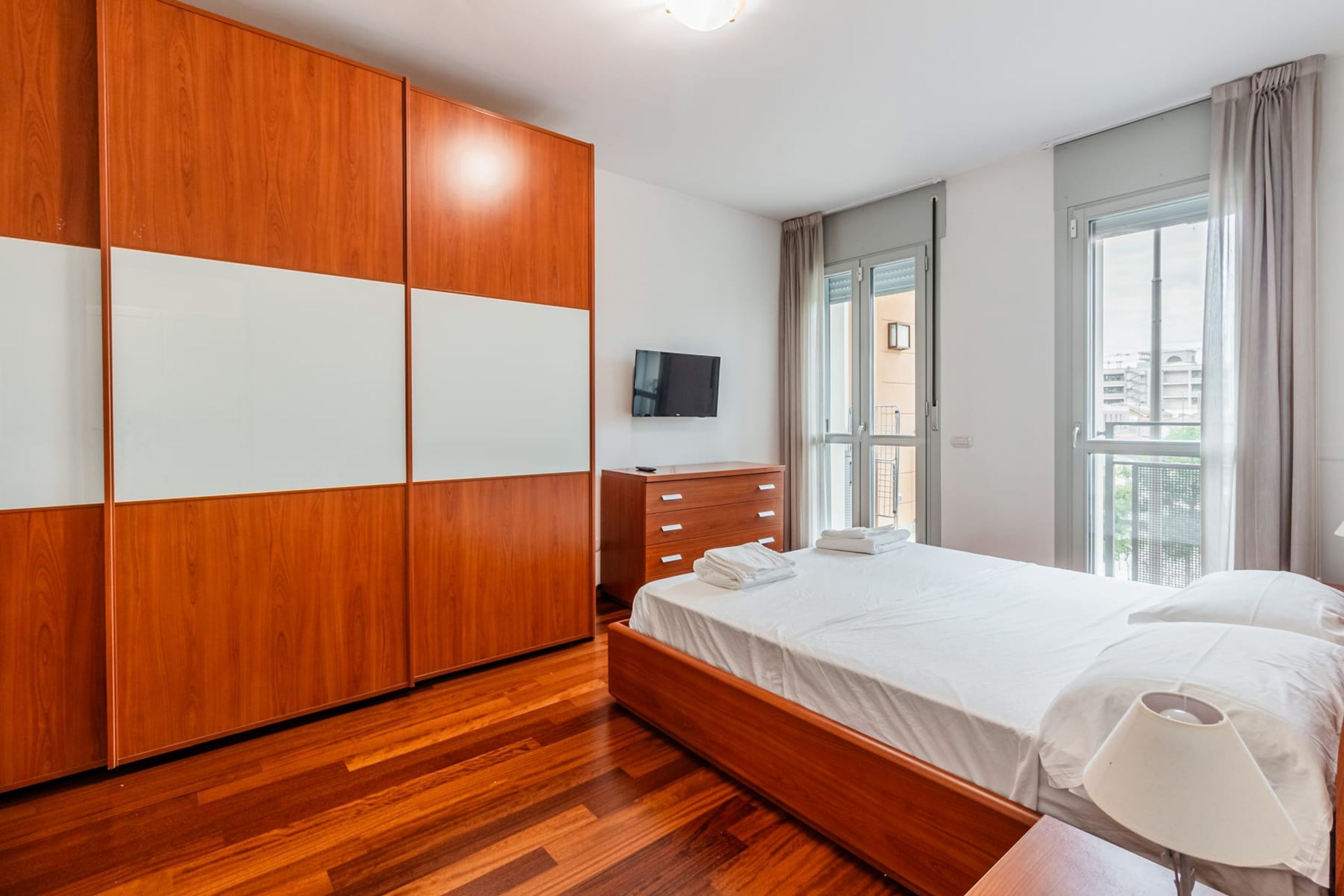 A modern bedroom with a wooden wardrobe, a bed with white linens, a bedside table, and a TV on the wall.