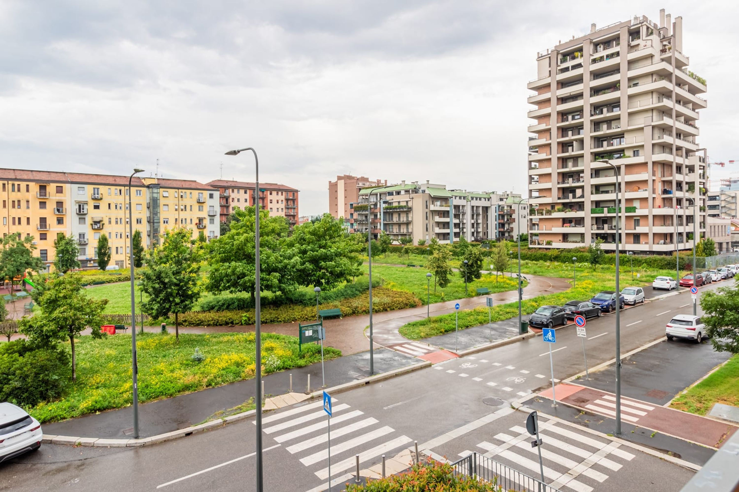 A multi-story residential building with balconies, surrounded by greenery and a pedestrian crossing.