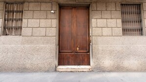 En trædør med metalgreb, mellem to vinduer med metalgitter, i en bygning med en struktureret facade.