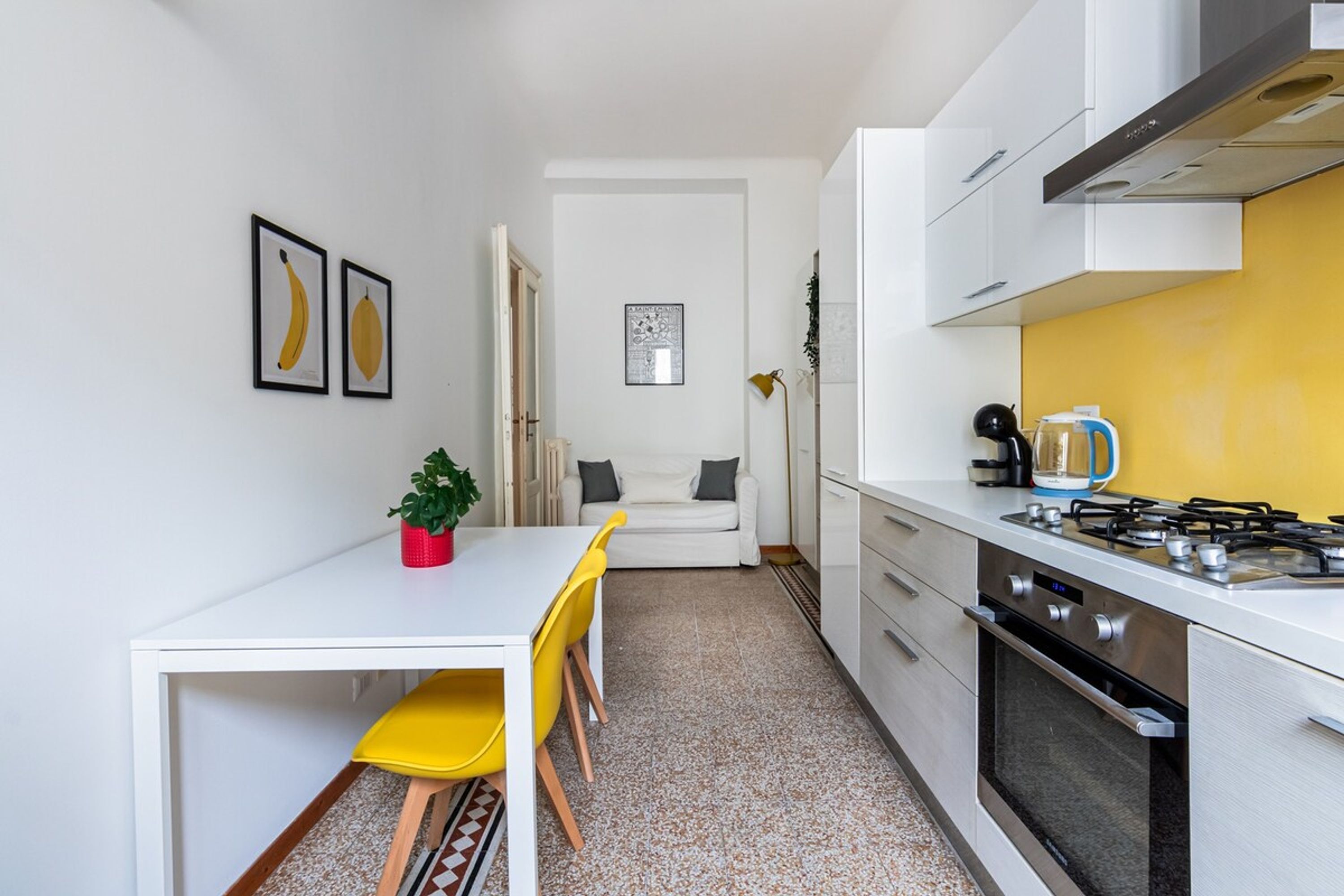 Een kleine keuken met een wit aanrecht, gele stoelen, een eettafel en een fornuis.