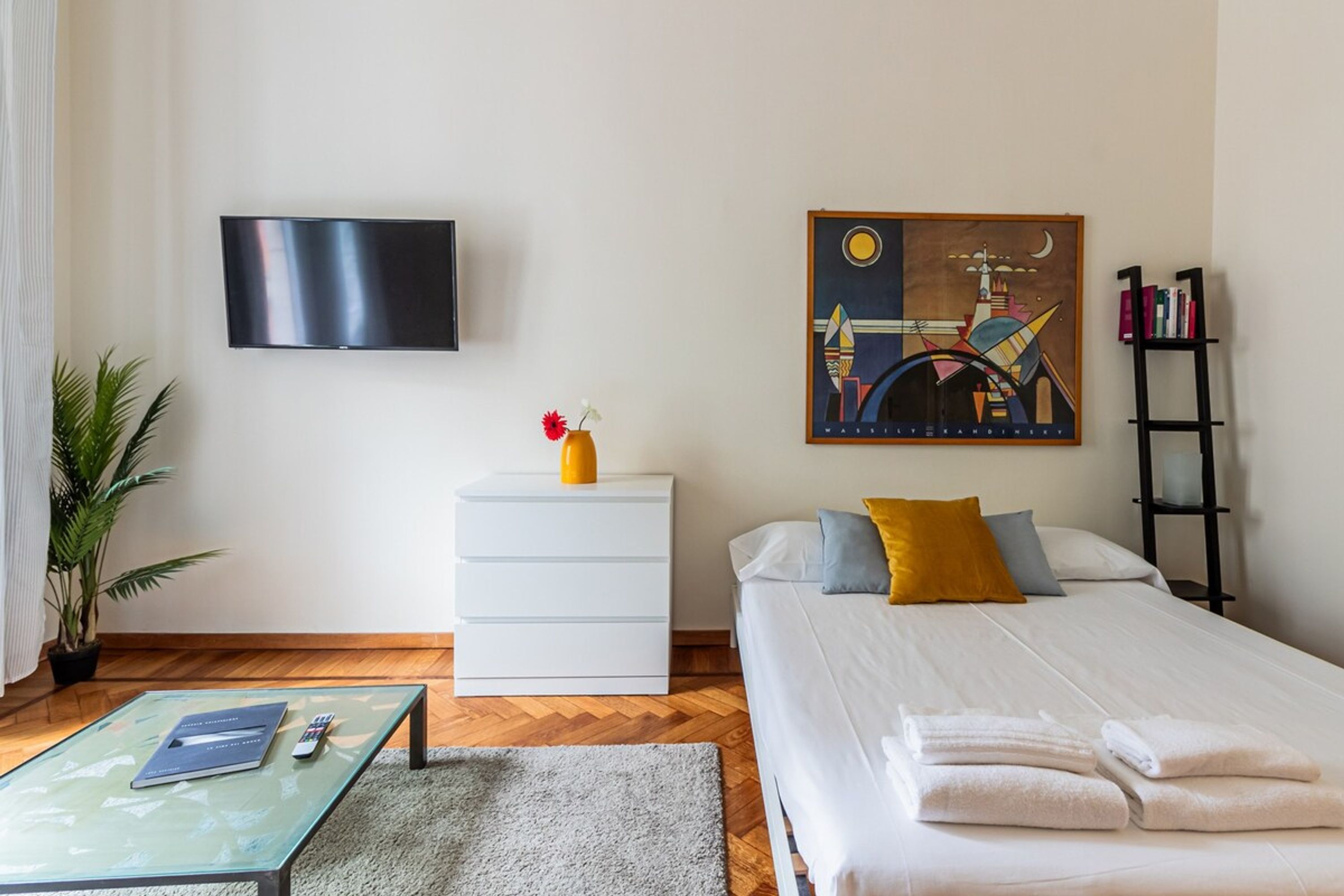 A hotel room with a bed, a flat-screen TV, a painting, a nightstand, a bookshelf, a coffee table, and a potted plant.