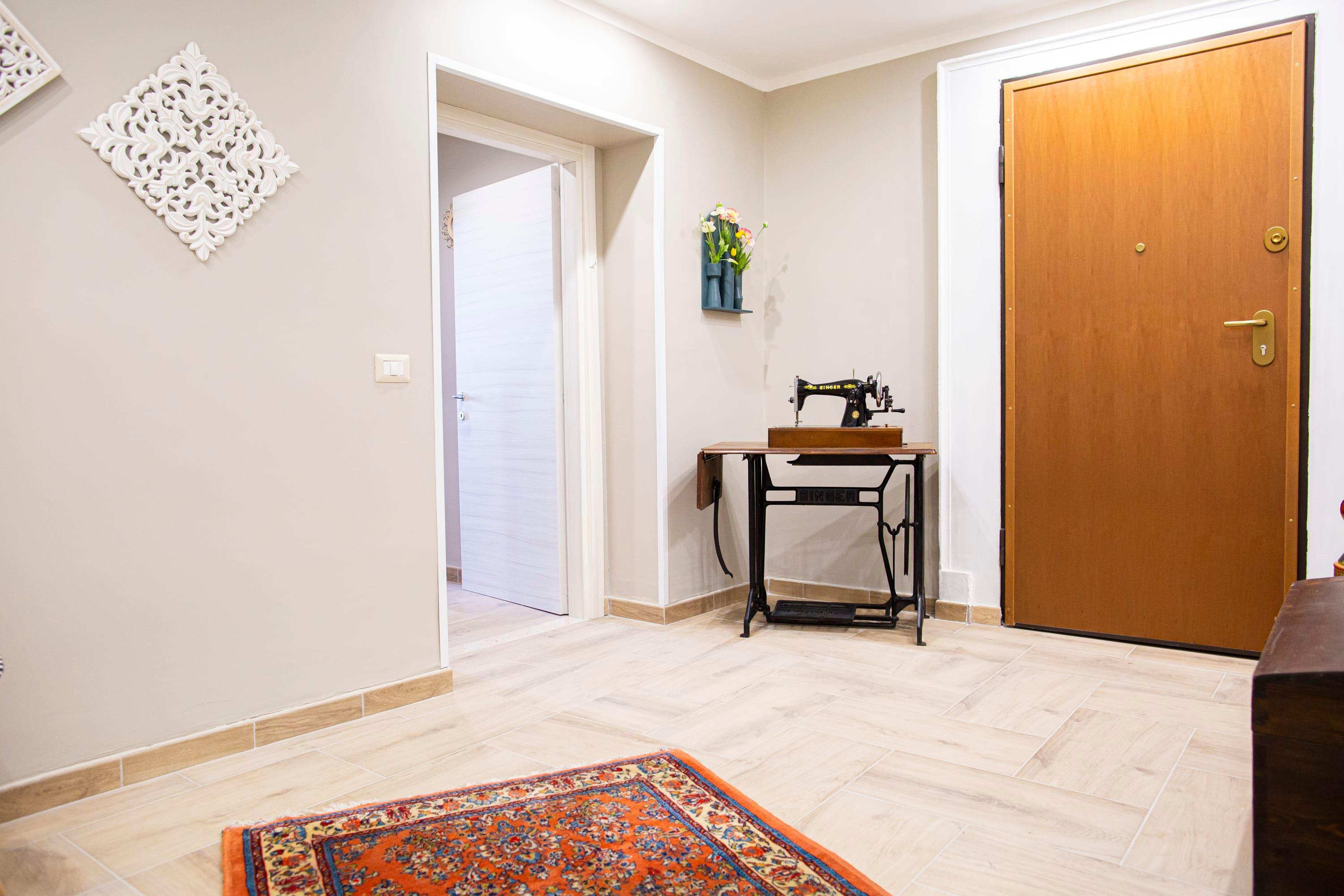Una habitación con puerta de madera, una máquina de coser sobre una mesa, un tapete estampado y un adorno en la pared.