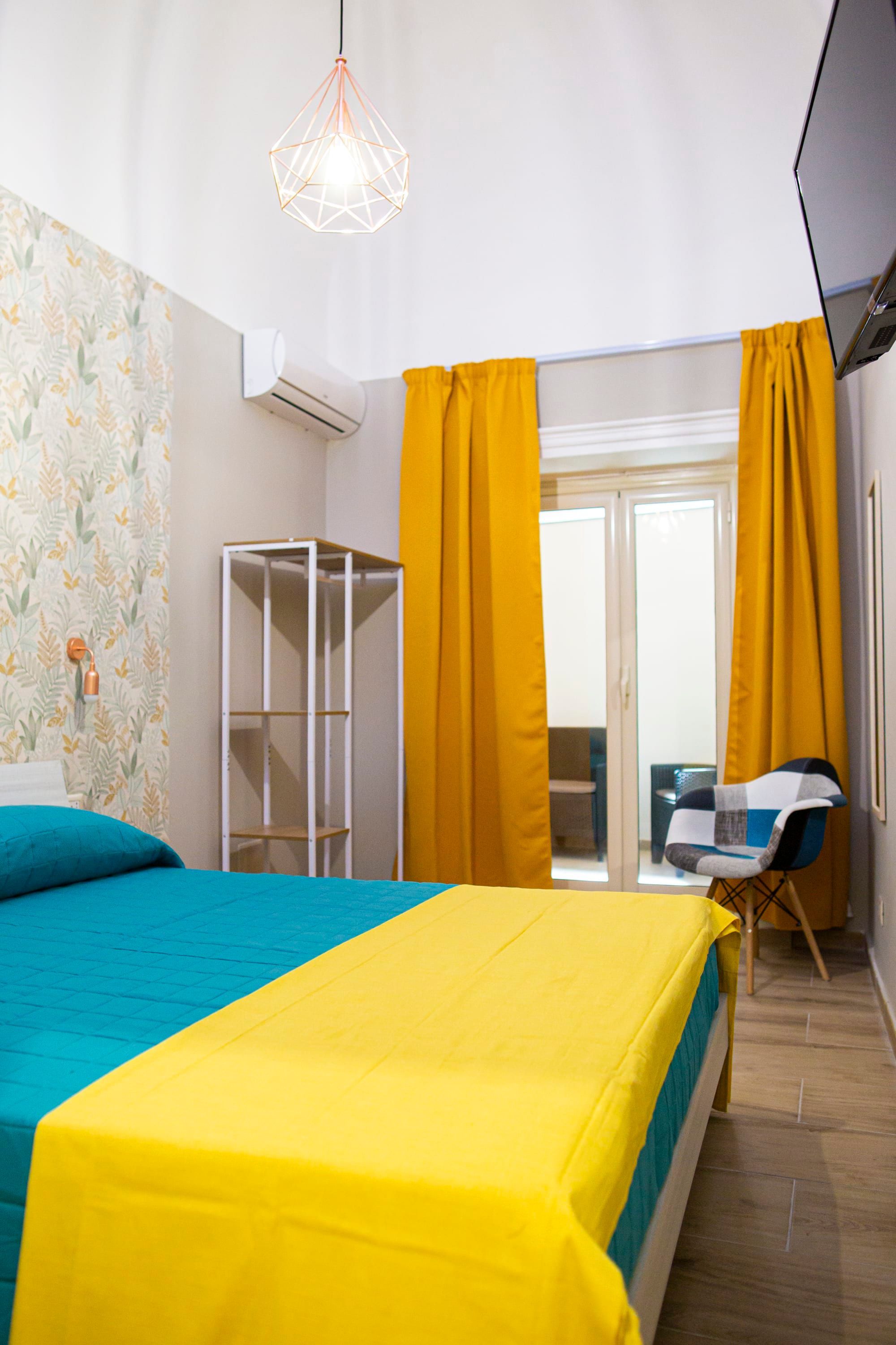 A bedroom with a bed, a wardrobe, a television, and a balcony with yellow curtains.