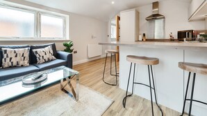 A modern living room with a glass coffee table, a sofa with patterned cushions, a kitchen with a stainless steel range hood, and wooden bar stools. - London Street Apt By REDWOOD STAYS (Basingstoke)
