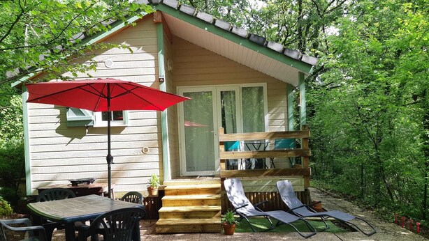 Eine kleine Holzhütte mit einer Terrasse, einem Tisch mit Stühlen, einem roten Sonnenschirm und zwei Liegestühlen.