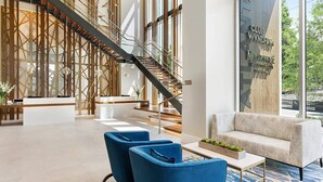 Una moderna hall d'albergo con una reception, poltrone blu, una scala e un'ampia finestra che offre vista sull'esterno.