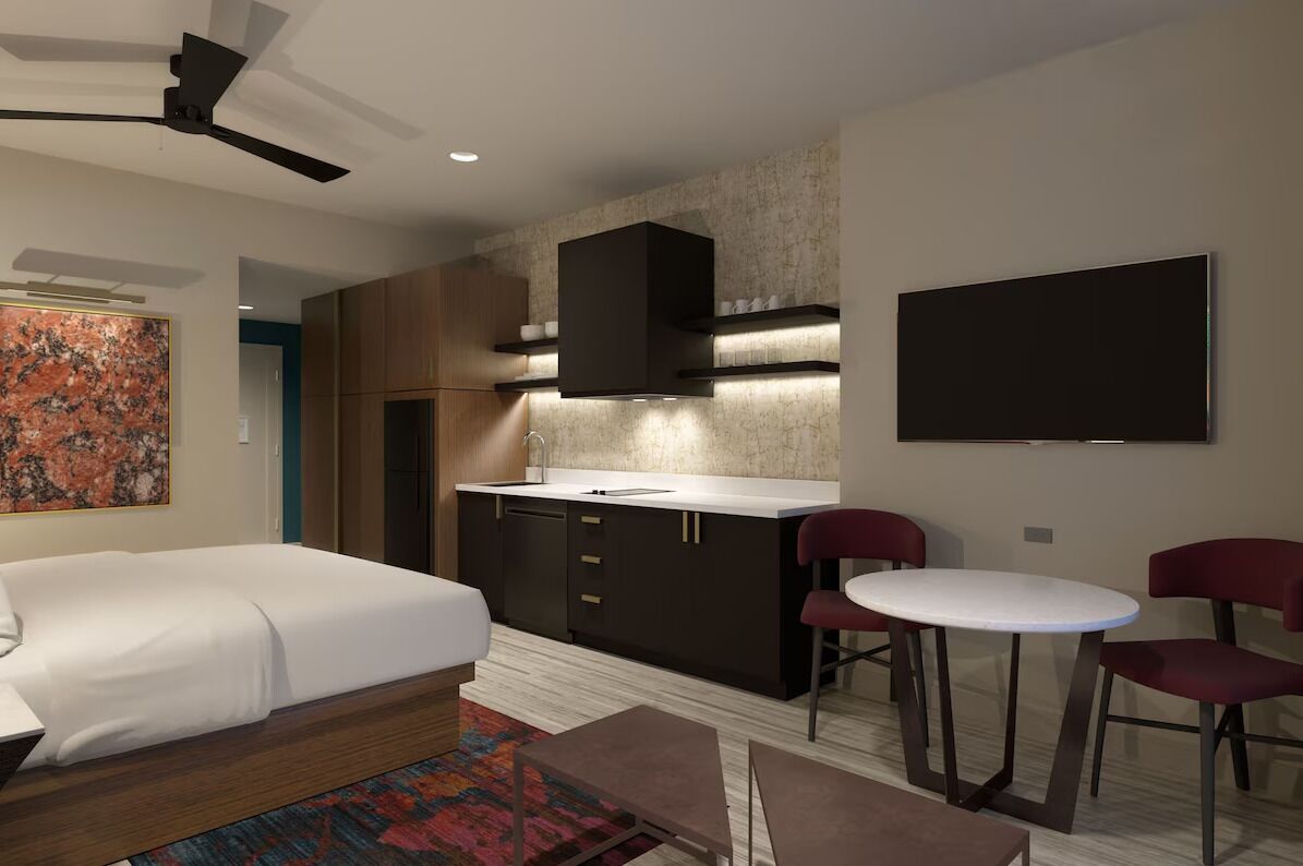 A modern hotel room with a bed, a kitchenette, a dining area with two chairs, and a large artwork on the wall.