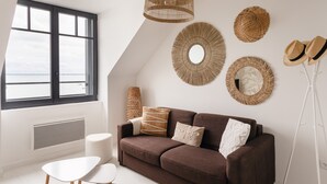 A modern living room with a brown sofa, white side table, and a large window with a view. - Ty Peskaiden 2 - Appt vue mer à Cancale (Cancale)