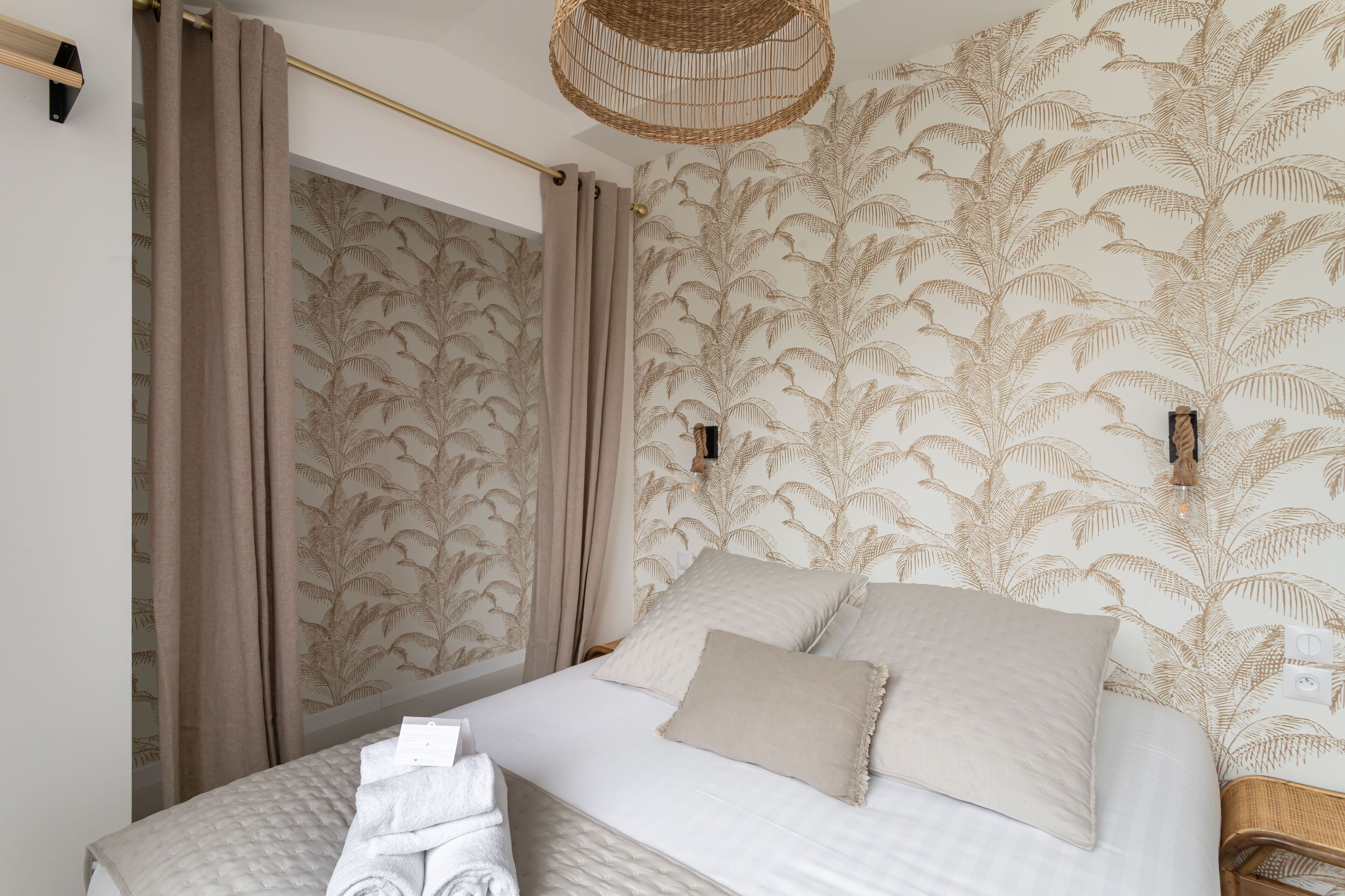 Un lit bien fait, recouvert d’une courtepointe, une table de chevet avec une lampe, et un mur orné d’un papier peint à motifs de feuilles.