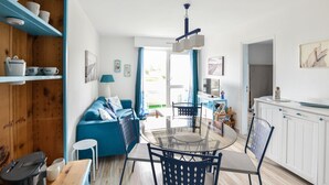 Una sala luminosa con un sofá azul, una mesa de comedor de vidrio y una cocina con armarios blancos.