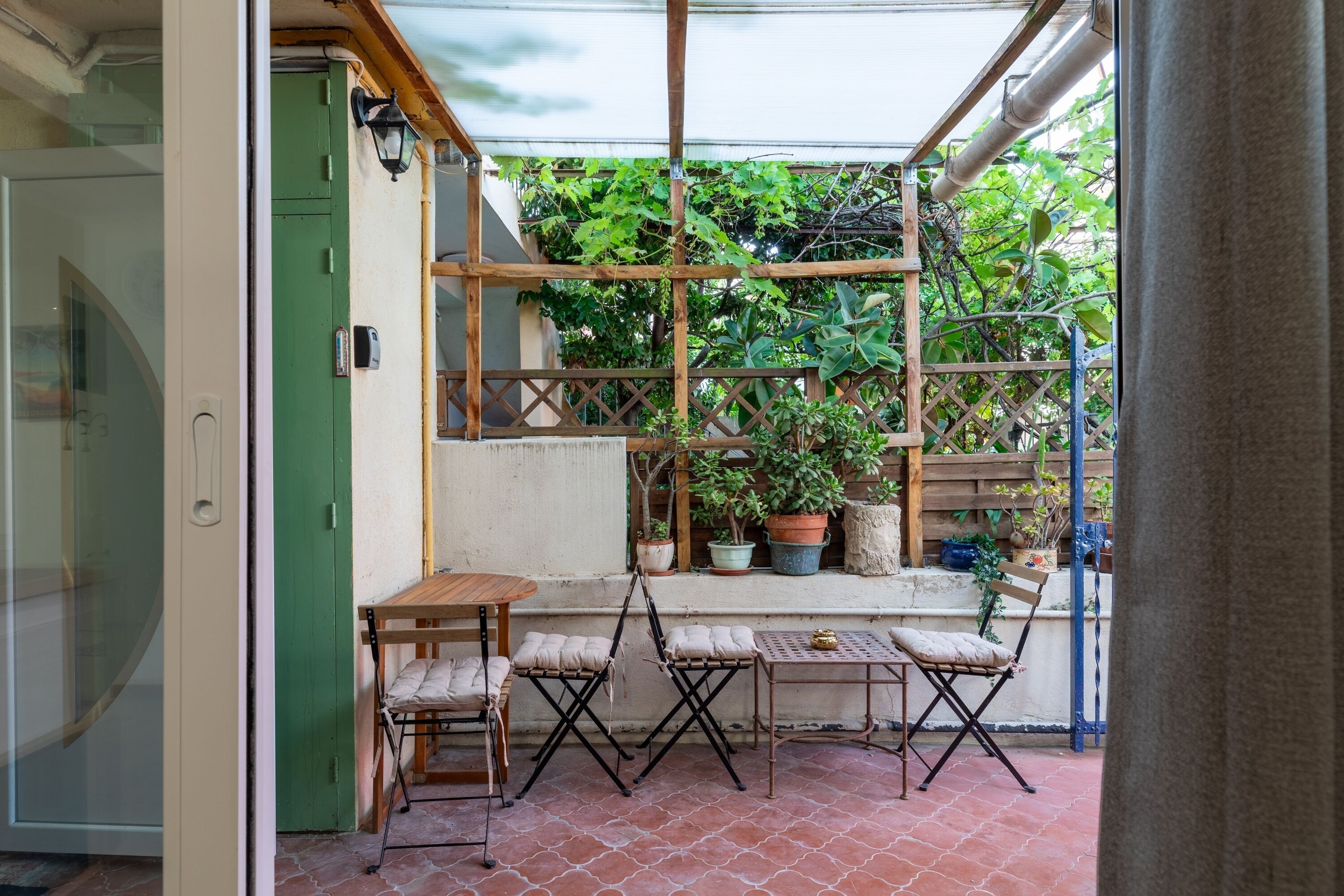 A patio area with a wooden table and chairs, a small table with a cushion, and potted plants.