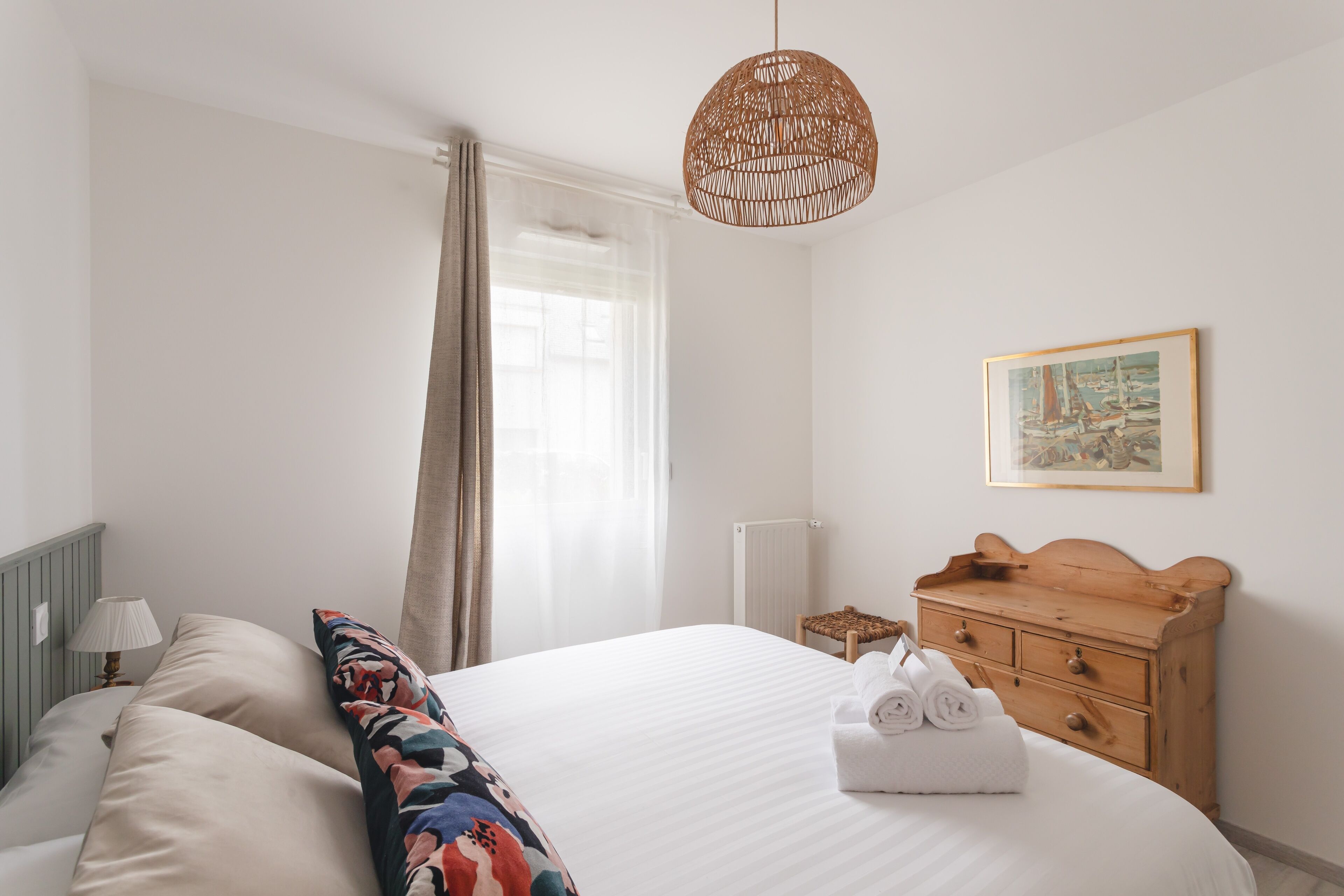 A neatly arranged bedroom with a large bed, a wooden dresser, a wicker hanging light, and a framed picture on the wall.