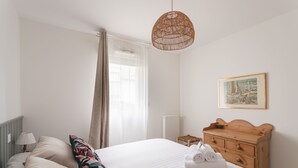 A neatly arranged bedroom with a large bed, a wooden dresser, a wicker hanging light, and a framed picture on the wall. - Les Sternes - Appt proche de la plage (Saint-Malo)
