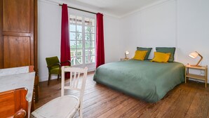 Ein Schlafzimmer mit Holzboden, einem grünen Bett mit gelben Kissen, einem Holzstuhl, einem Holztisch und einem Fenster mit roten Vorhängen.