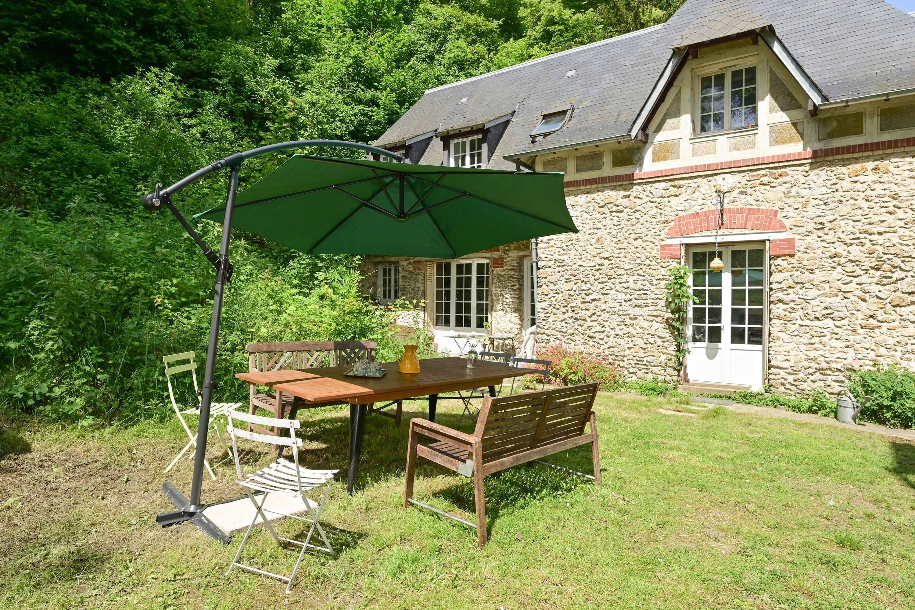 Een eethoek buiten met een tafel en stoelen, een groene parasol en een stenen gebouw op de achtergrond.