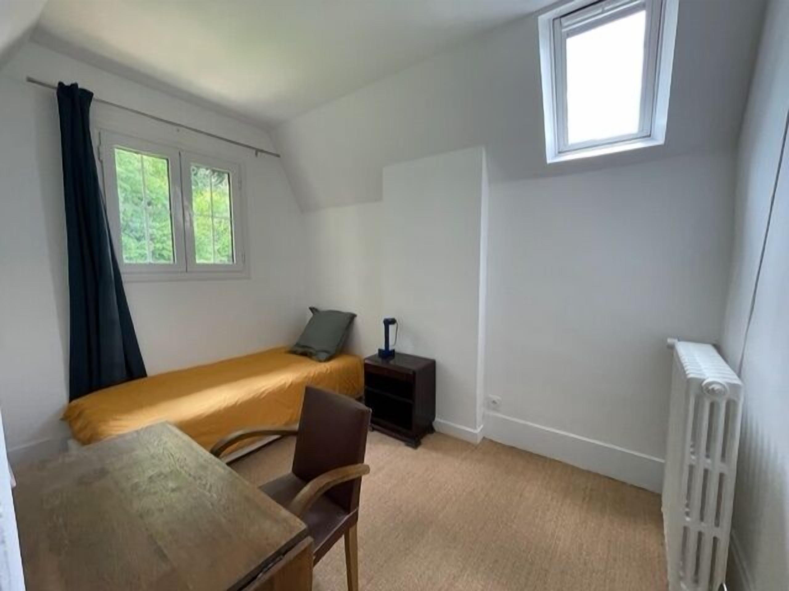 A small, sparsely furnished room with a single bed, a desk, and a chair. There are two windows with curtains, a radiator, and a small table with a lamp.