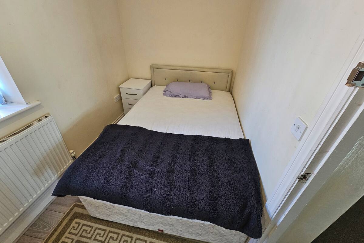 A small, single-bed room with a beige headboard, a white bedside table, and a window with blinds.
