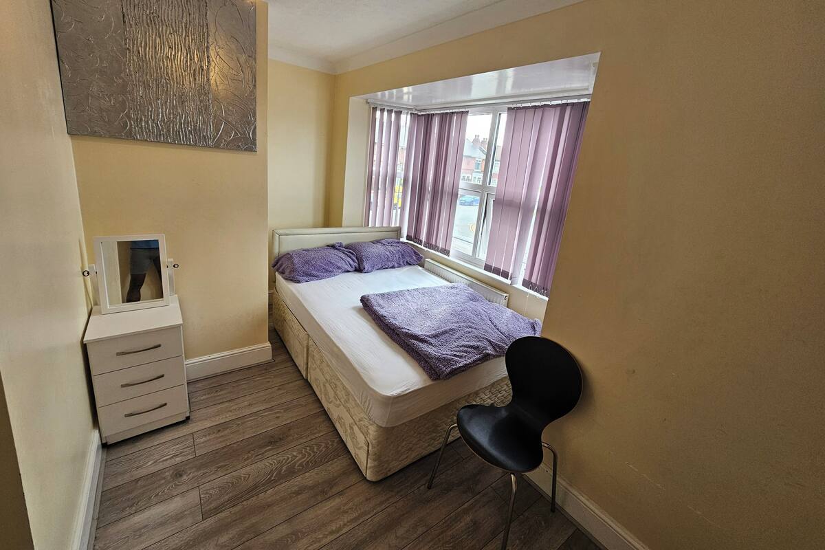 A bedroom with a bed, a bedside table, a chair, a window with curtains, and a wall-mounted artwork.