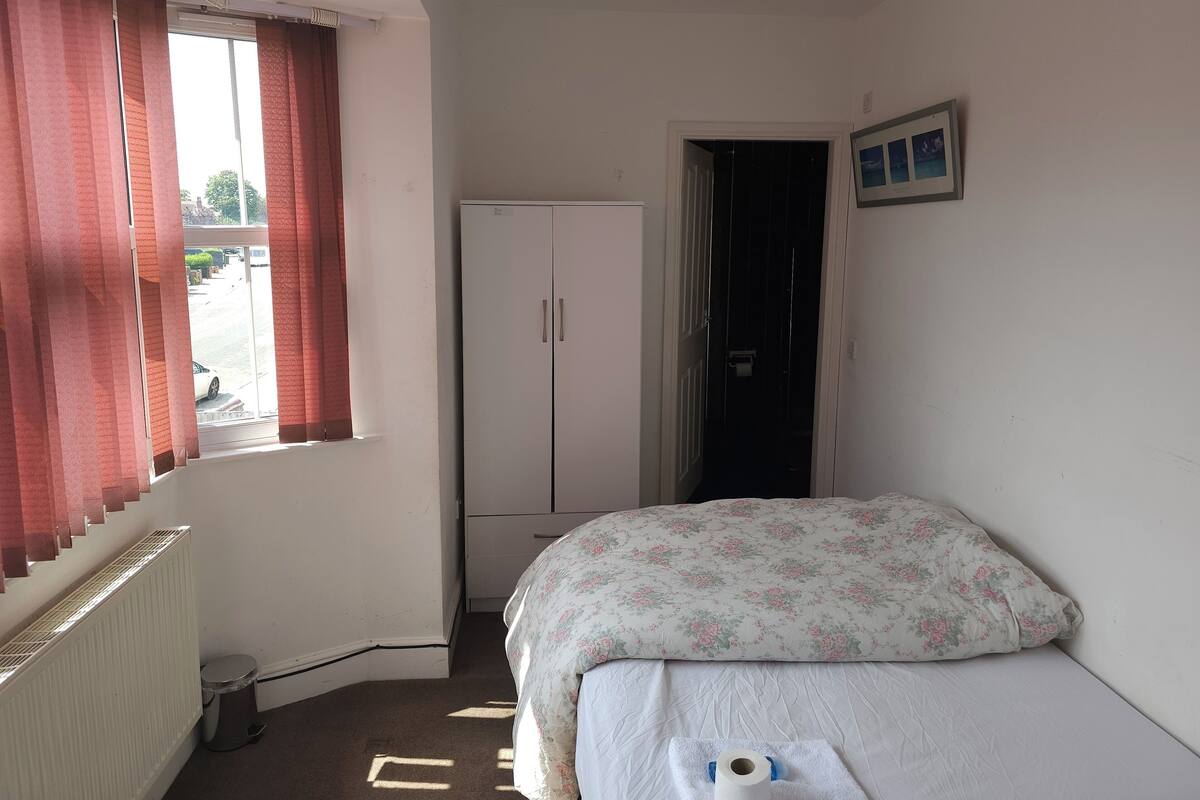 A small, sparsely furnished room with a bed, a radiator, a wardrobe, and a window with curtains.