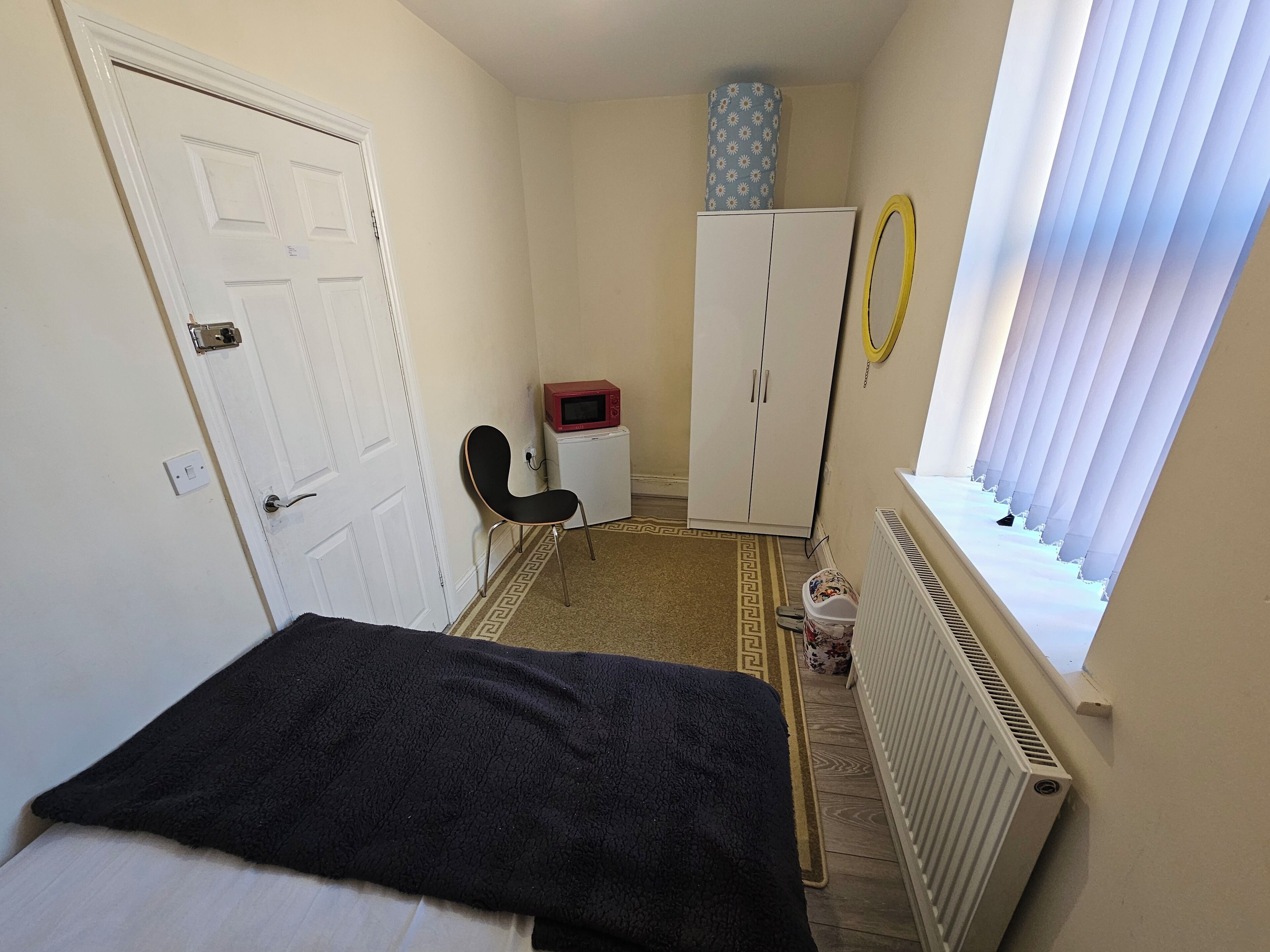A small, narrow room with a bed, a chair, a wardrobe, a radiator, a window with blinds, and a small table with a red object on it.