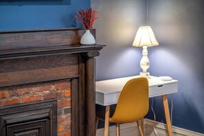 Ein Zimmer mit Kamin aus Ziegelsteinen, einem weißen Schreibtisch, einem senffarbenen Sessel und einer Lampe.