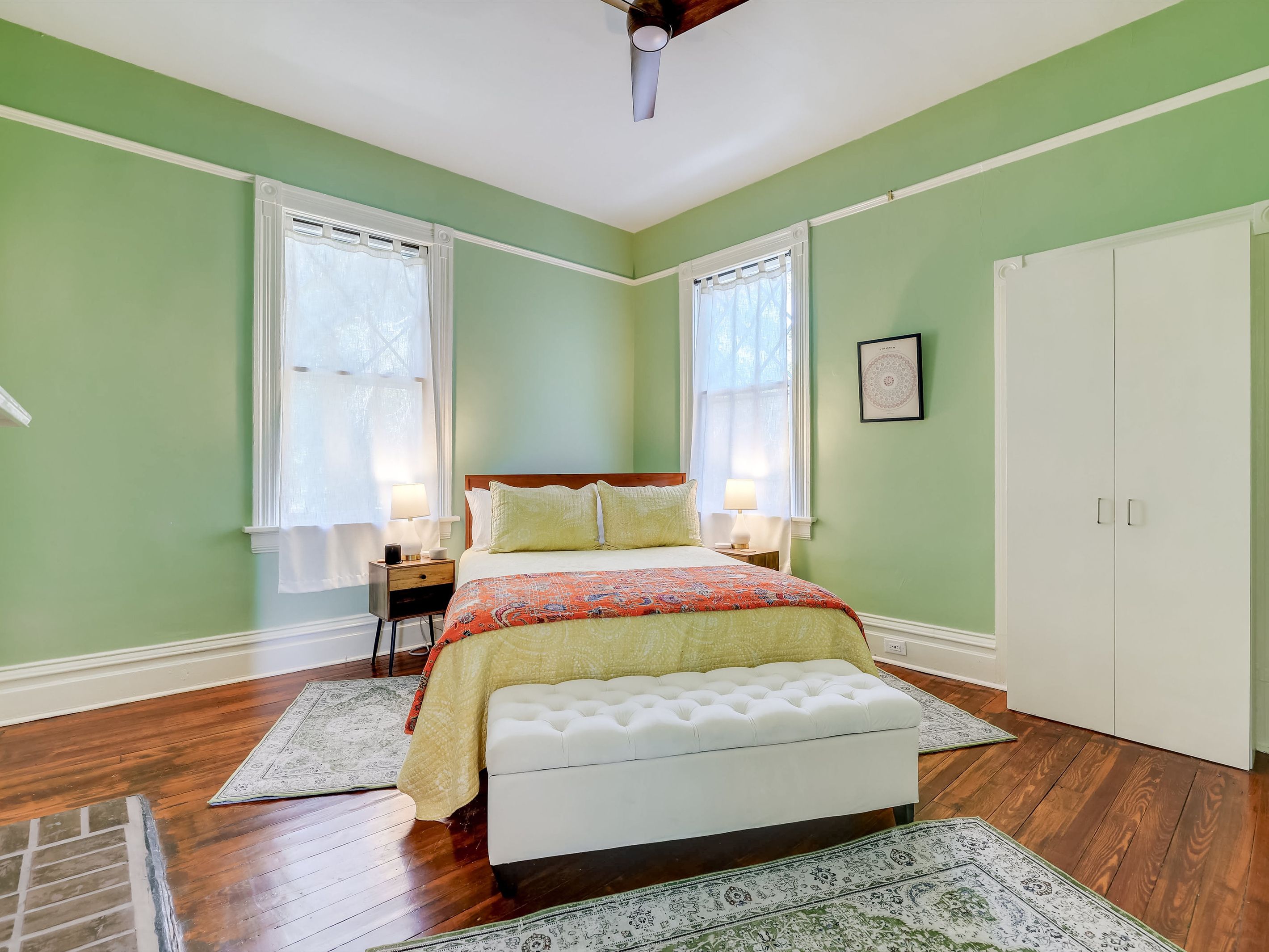 Une chambre à coucher comprenant un lit, des tables de chevet, un banc, une armoire et deux fenêtres avec des rideaux.