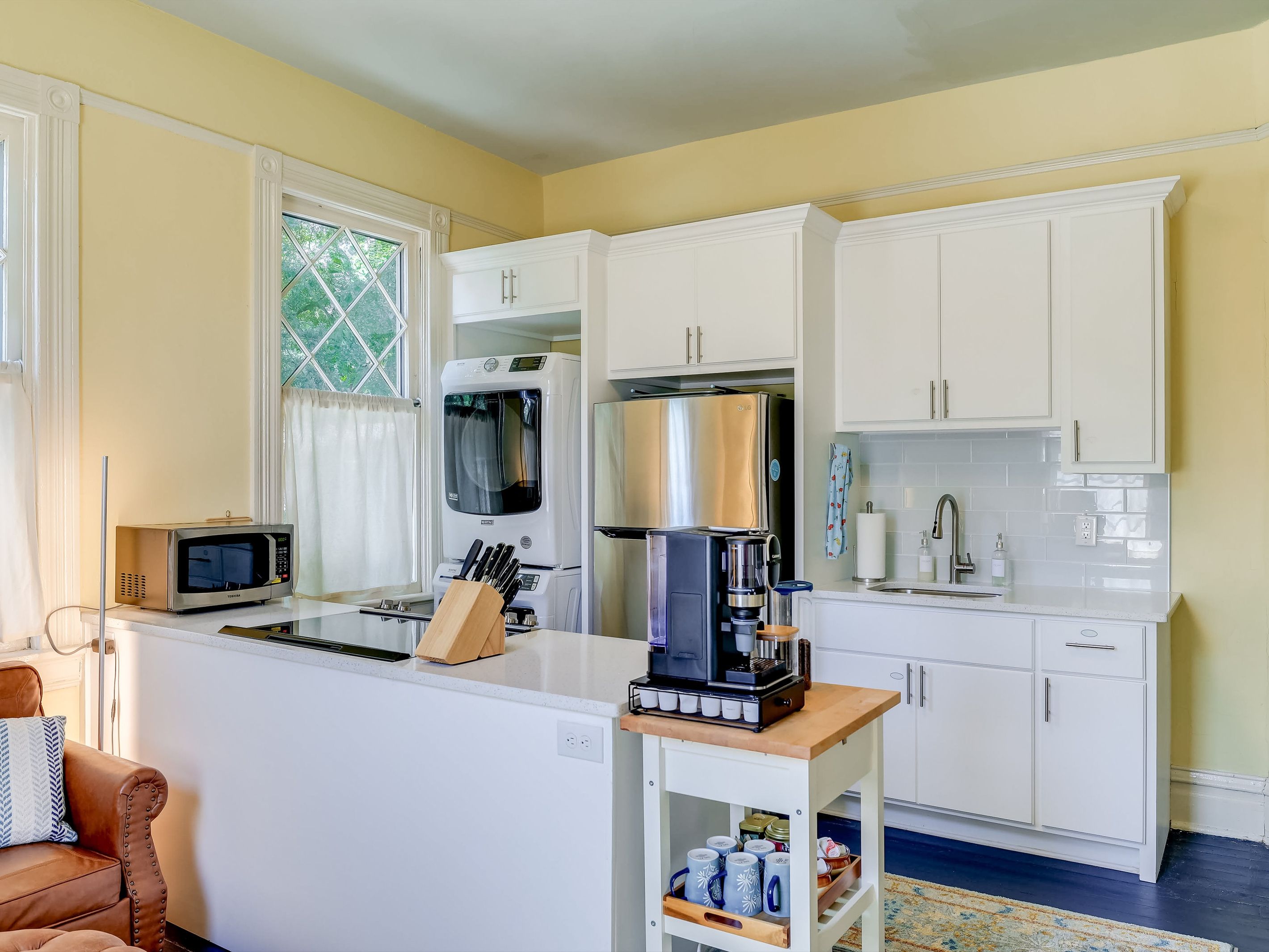 A compact kitchen with white cabinetry, a coffee machine, a microwave, and a window with curtains.