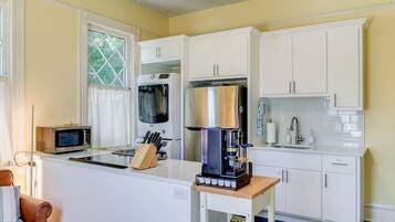 A compact kitchen with white cabinetry, a coffee machine, a microwave, and a window with curtains.