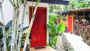 Une maison en bois avec une porte rouge, entourée d’une végétation luxuriante et de plantes en pot.