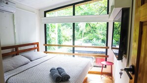 Une chambre avec un grand lit, une tête de lit en bois, une fenêtre donnant sur un paysage verdoyant et un tabouret rouge.