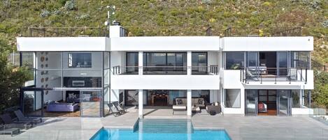Una casa moderna de dos pisos con piscina, mobiliario exterior y vista a un paisaje montañoso.