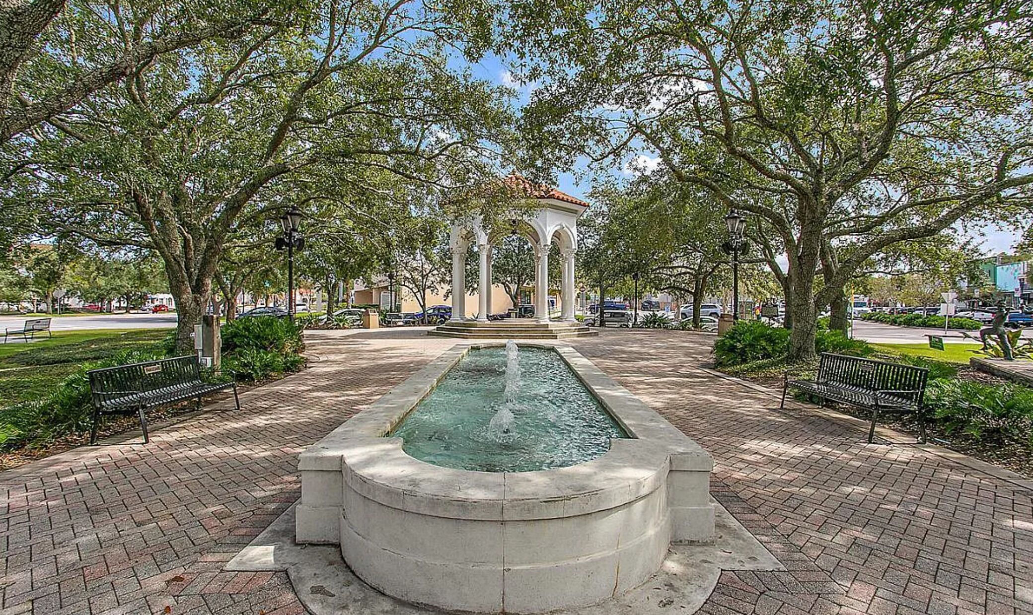 A park with a central fountain, surrounded by trees and benches.