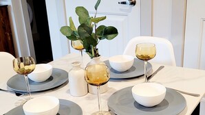 A dining table set with white bowls, grey plates, and wine glasses, accompanied by a vase of green leaves and a salt and pepper shaker.