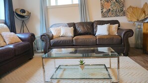 A living room with a brown leather sofa, a glass coffee table, and a large window with curtains.