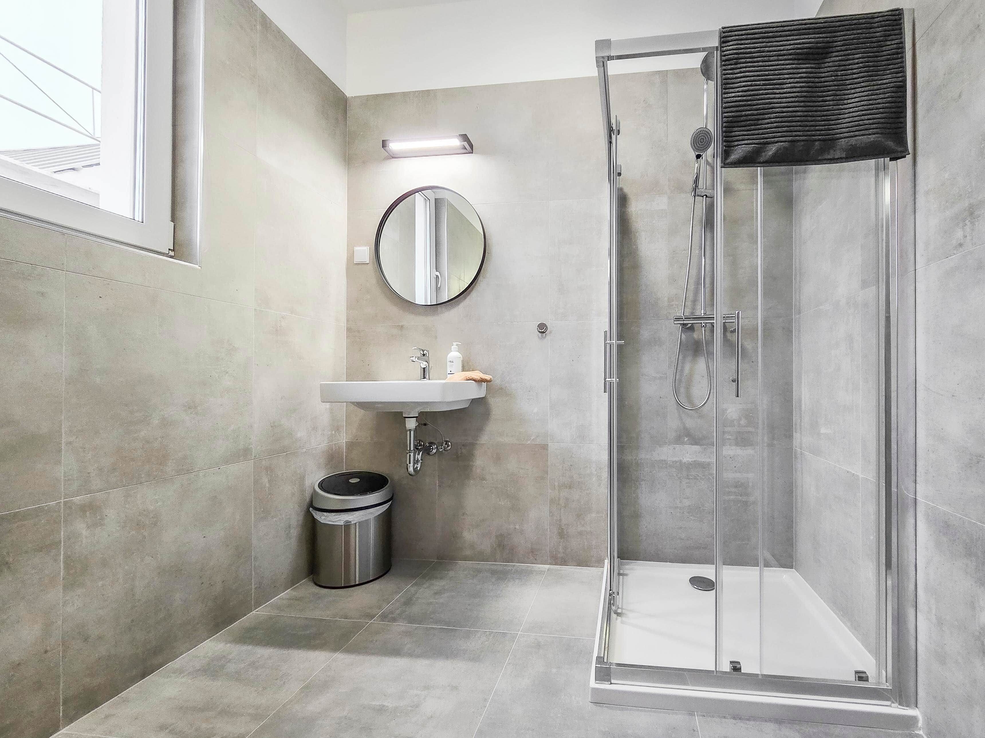 A modern bathroom with a glass-enclosed shower, a sink with a mirror above it, and a trash bin.