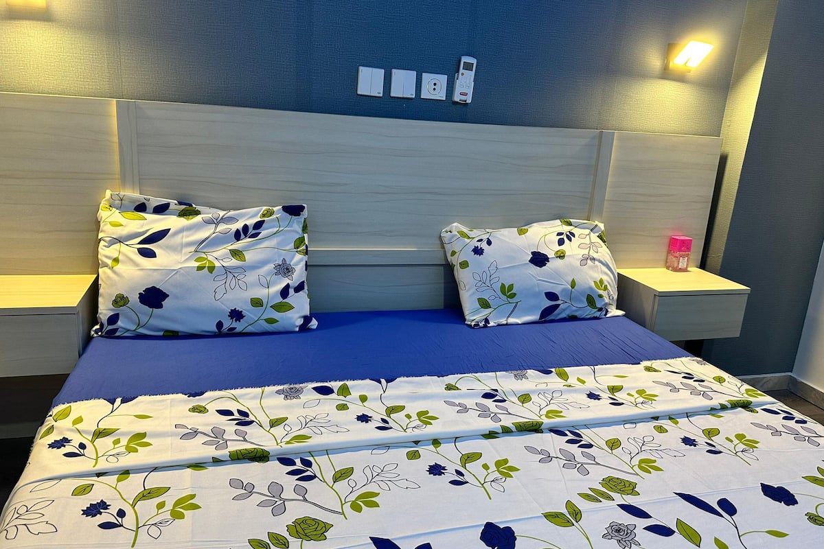 A neatly made bed with a floral bedspread and two matching pillows, flanked by nightstands with a small lamp and a bottle.