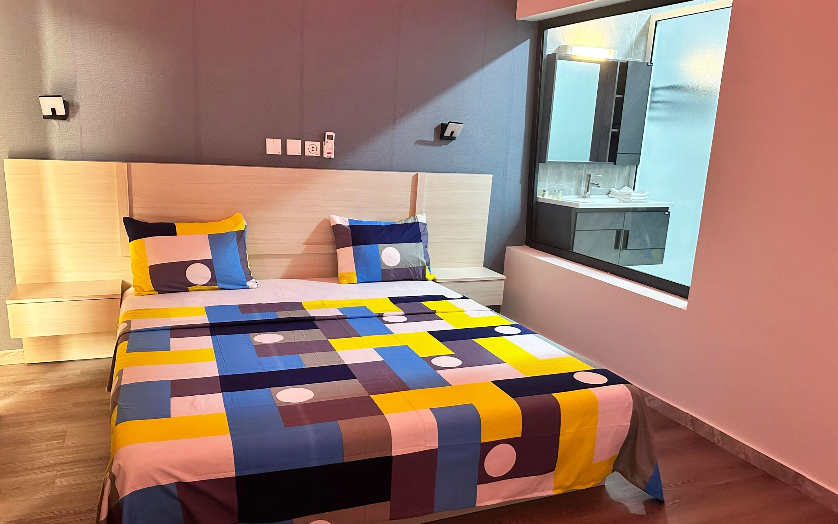 Un dormitorio moderno con una cama grande, un cabecero, una ventana con vistas a un baño y un interruptor de luz en la pared.