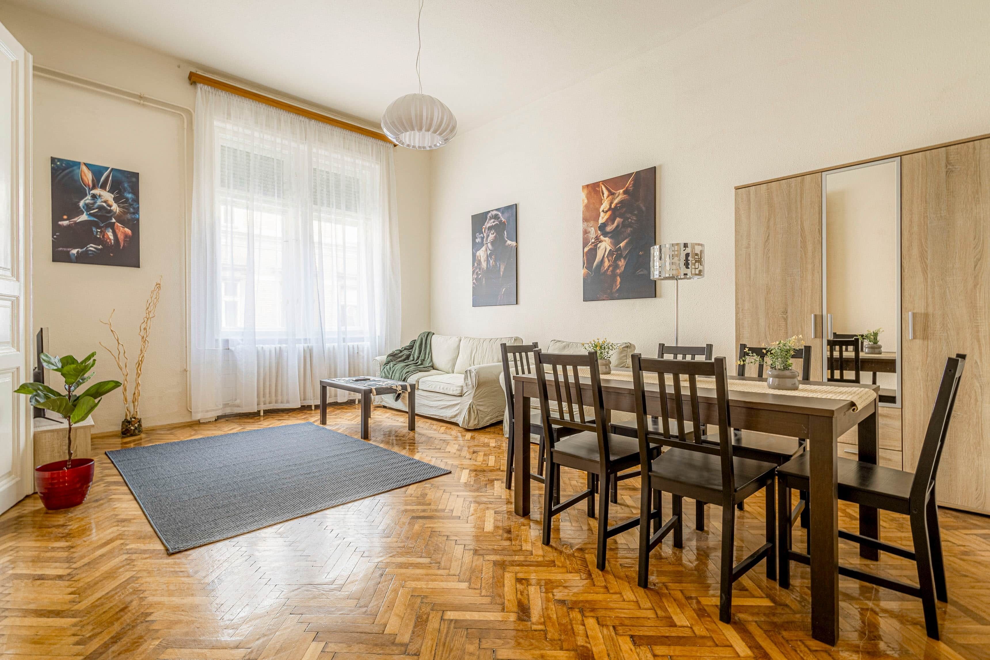 A spacious living area with a wooden herringbone floor, a dining table with chairs, a sofa, and a large window with sheer curtains.