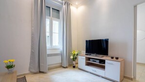 Een moderne woonkamer met een flatscreen televisie op een houten kast, een raam met gordijnen, en een kleine tafel met een bloemenvaas.
