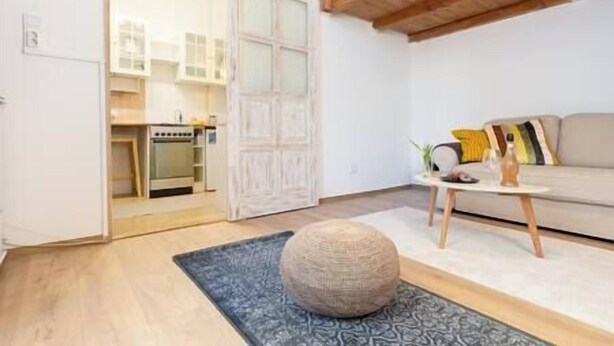 A modern living room with a round ottoman, a sofa with cushions, a small table with a vase, and a kitchen visible through an open door.