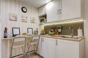 A modern kitchen with white cabinets, a stainless steel sink, and a microwave above the counter. There are two bar stools and a clock on the wall.