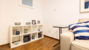 A room with a white wall, a framed picture, a small table with a black frame, a beige armchair with a blue and white striped pillow, and a white shelving unit with various decorative items. - IMCT - Beautiful Home at Basilica 1 (Budapest)