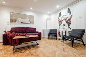 A modern living room with a herringbone wooden floor, a red sofa, two black chairs, a small wooden coffee table, and a wall-mounted mirror.